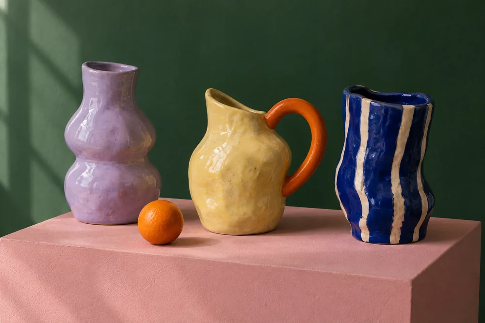 Three ceramic vessels in lilac, yellow and blue on a pink plinth against a green backdrop.